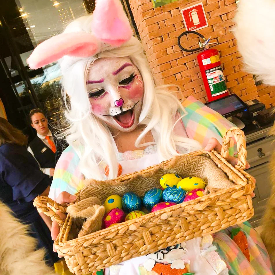 Um Coelho Laranja animado liderando uma caça aos ovos de Páscoa com várias crianças correndo e sorrindo atrás dele em um parque